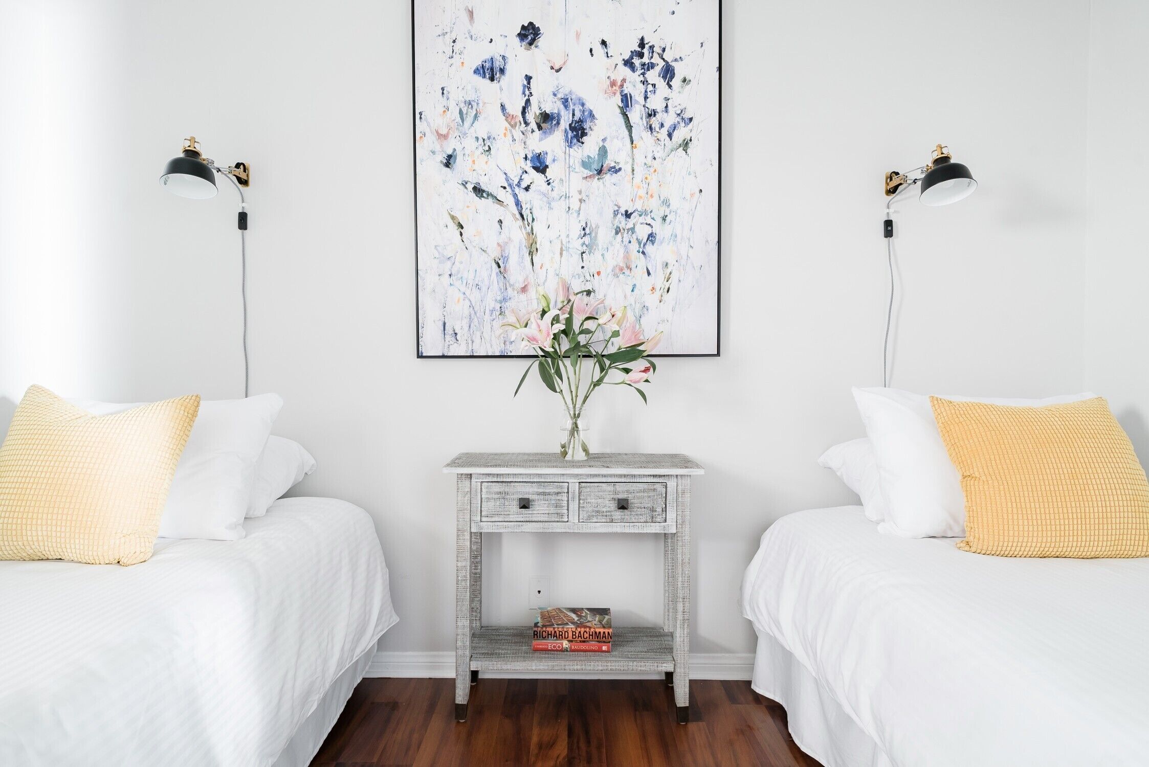 A bedroom with two beds, a bedside table with a lamp, a framed floral artwork, and a vase with flowers.