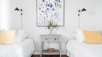 A bedroom with two beds, a bedside table with a lamp, a framed floral artwork, and a vase with flowers.