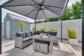 Un espace extérieur abrité, avec une table et des chaises, un petit abri de jardin et, au fond, une clôture blanche.