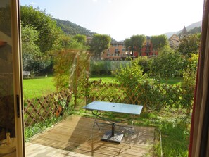Une vue depuis une fenêtre donnant sur un jardin agrémenté d’une terrasse en bois, d’une table et d’une pergola.