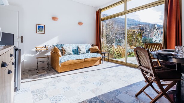 A modern living room with a sofa, dining table, and chairs, large windows with a view of a town and hills. - Le Garibaldi - Studio cosy Sospel (Sospel)