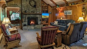 A cozy living room with a stone fireplace, wooden beams, a ceiling fan, and a television. - Williams House at Lake Junaluska -Spacious 5-Bedroom Family Retreat (Lake Junaluska)