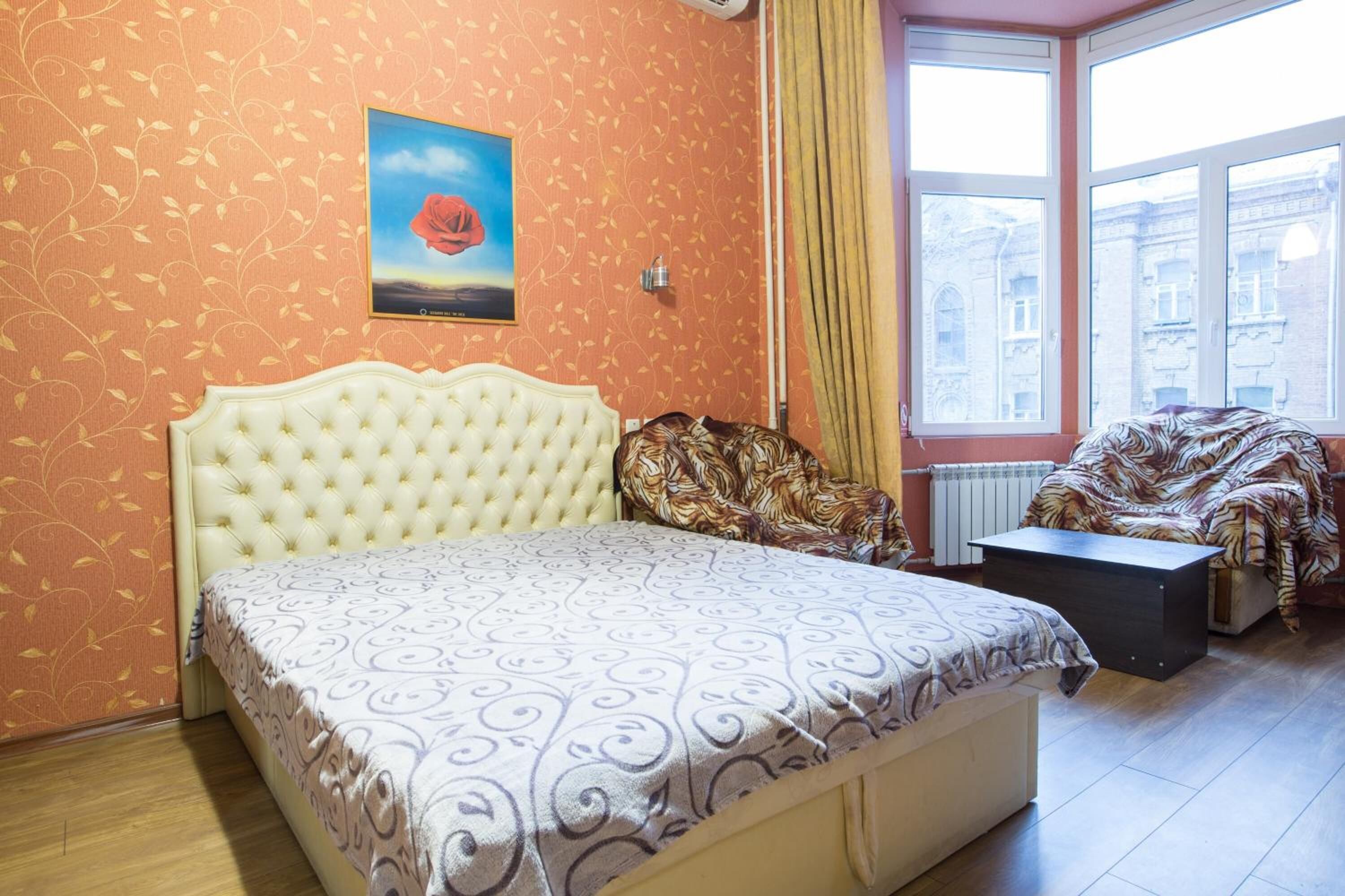 A bedroom with a tufted headboard, patterned bedspread, a framed picture on the wall, a window with curtains, and a view of buildings outside.