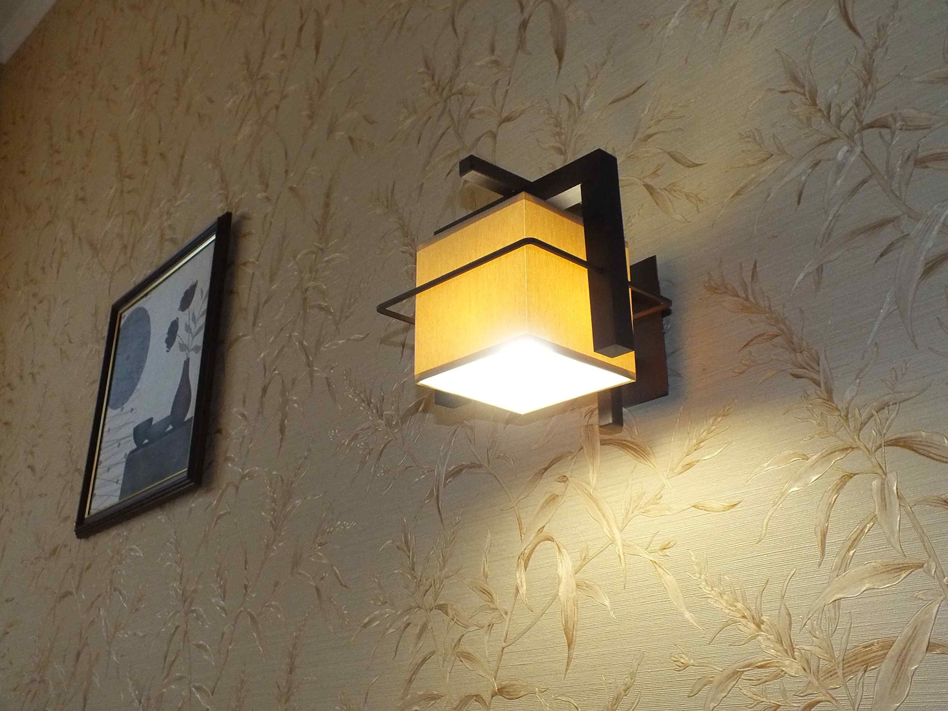 En väggmonterad lampa med ett geometriskt mönster, en inramad tavla och en vägg med en tydlig struktur.