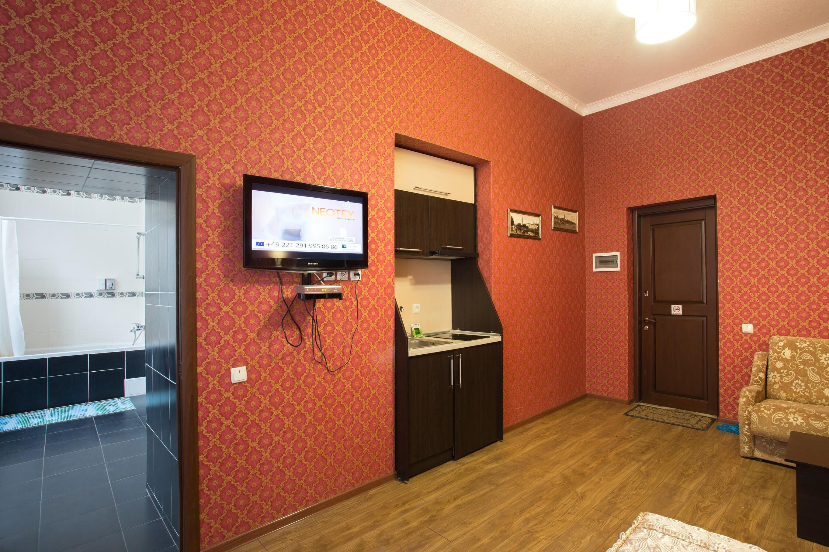 A room with a television mounted on the wall, a small kitchen area with cabinets, a door, and a patterned wall.