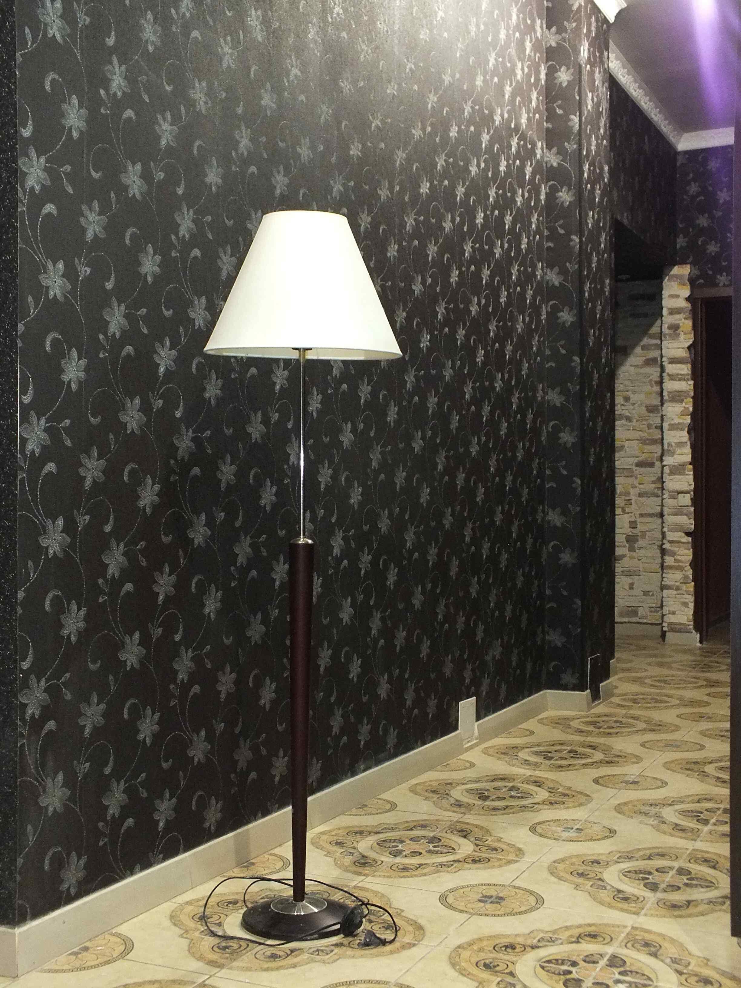 Eine Stehlampe mit weißem Lampenschirm in einer Diele mit strukturiertem Wandpapier und Fliesenspiegel.