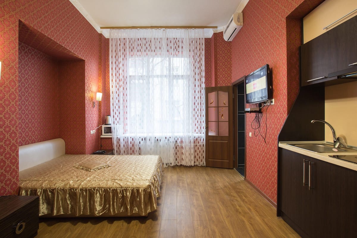 Quarto com cama, mesa de cabeceira, televisão, janela com cortinas e cozinha compacta com pia.