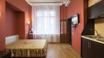 Un dormitorio con cama, mesita de noche, televisor, ventana con cortinas y kitchenette con lavabo.