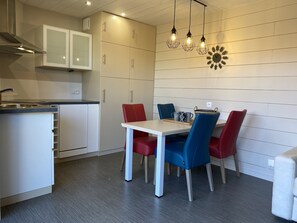 A modern kitchen with white cabinets, a dining table with red and blue chairs, and pendant lights above.