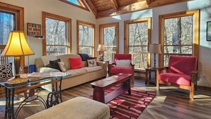 A spacious living room with a wooden ceiling, large windows, a sofa, chairs, a coffee table, and a lamp.