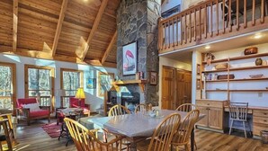 A spacious living area with a high ceiling, wooden flooring, a dining table with chairs, a fireplace, and a stone wall.