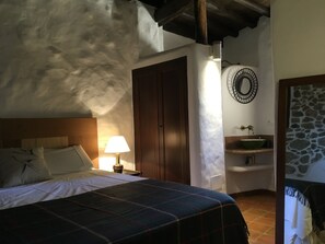 Une chambre à coucher comprenant un lit, une table de chevet, une lampe, une porte en bois, un mur en pierre et un élément décoratif circulaire fixé au mur.