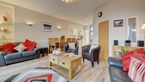 Un salon avec un canapé, une table basse et des fauteuils. On y trouve également une table à manger avec des chaises, un coin cuisine et une porte qui donne sur une autre pièce.