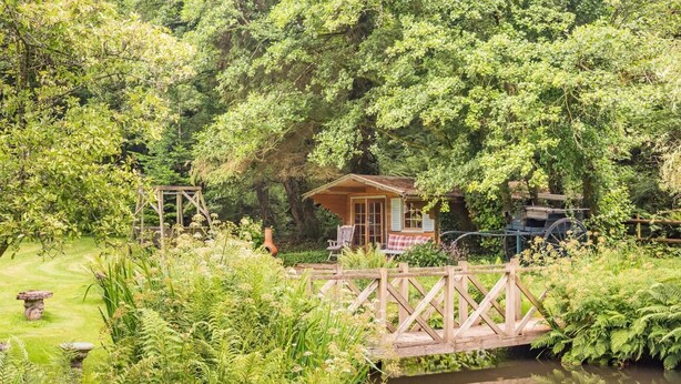 A rustic wooden bridge over a pond, surrounded by lush greenery and a small wooden cabin with a porch. - 1 Bed in East Down (oc-kincot) (East Down)