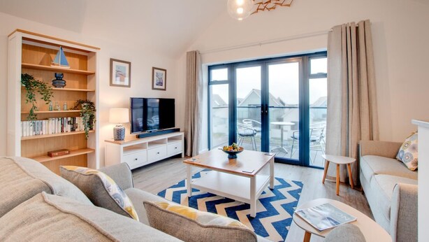 A modern living room with a flat-screen TV, a bookshelf, a dining area, and a large sliding glass door leading to an outdoor patio. - 2 Bed in Westward Ho! (oc-idleho) (Northam)