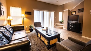 A living room with a leather sofa, a glass coffee table, a TV, and a staircase leading to another level. - Cozy - Adorable 3 Bed 2.5 Bath Townhouse 1015 (Kissimmee)