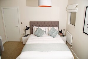 A neatly arranged bedroom with a tufted headboard, a bed with white bedding, two bedside lamps, and a window with a blind.