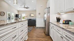 A modern kitchen with white cabinetry, a stainless steel refrigerator, and a granite countertop. - Glide Path: Dog-Friendly, Pool, Lift, Near Dock (Oak Island)