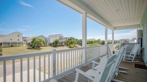 A covered porch with white rocking chairs, a wooden deck, and a view of a residential area with houses and greenery. - Glide Path: Dog-Friendly, Pool, Lift, Near Dock (Oak Island)