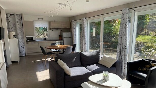 A modern living room with a dark grey sofa, a small round table, and a dining area with a wooden table and chairs. - Les Cottages du golf 3/3 (LE TRONCHET)