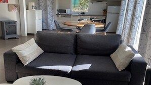 A modern living room with a dark grey sofa, a small white coffee table, and a dining area with a wooden table and chairs. - Les Cottages du golf 3/3 (LE TRONCHET)