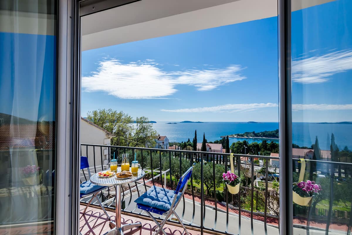 Un balcon avec une table préparée pour le petit-déjeuner, offrant une vue panoramique sur la mer et les collines.