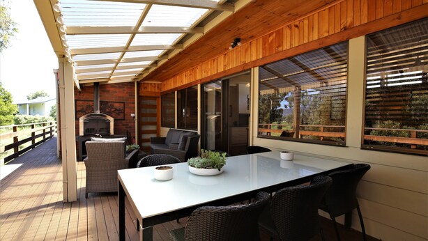 Een terras buiten met een tafel en stoelen, een open haard van baksteen en grote ramen.