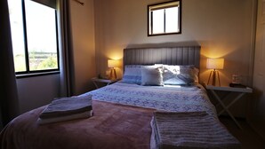 Een opgemaakt bed met een beddekleed met patroon, twee bedlampen en een raam met gordijnen.