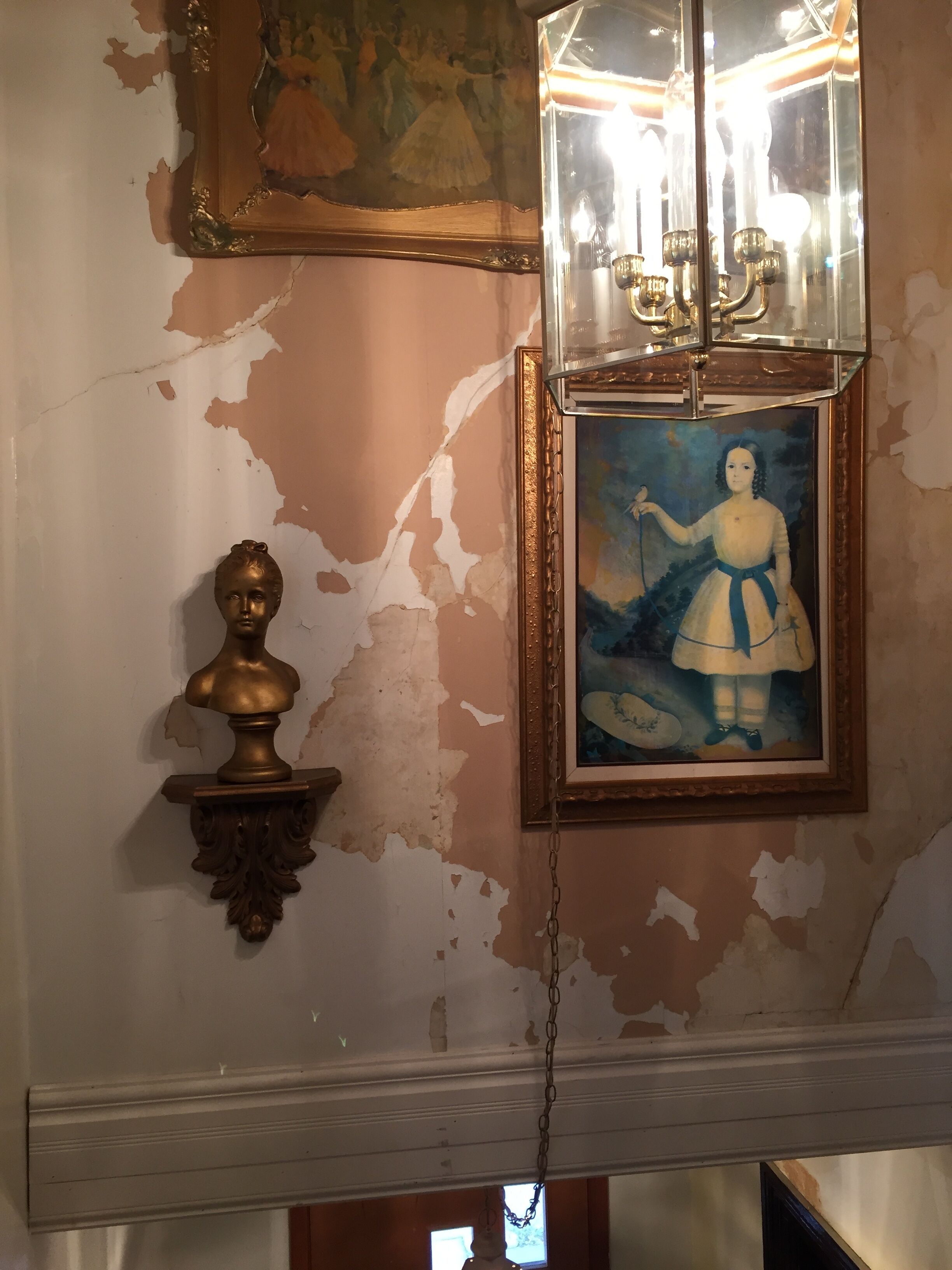 A room with peeling wall paint, a chandelier, a bust, and a framed painting.