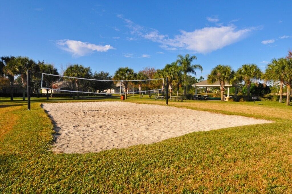 Ein Beachvolleyballfeld mit Netz, umgeben von Sand und Gras, mit Palmen und einer Laube im Hintergrund.