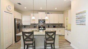 Una cucina moderna con mobili bianchi, piani in granito, elettrodomestici in acciaio inossidabile e illuminazione a sospensione.