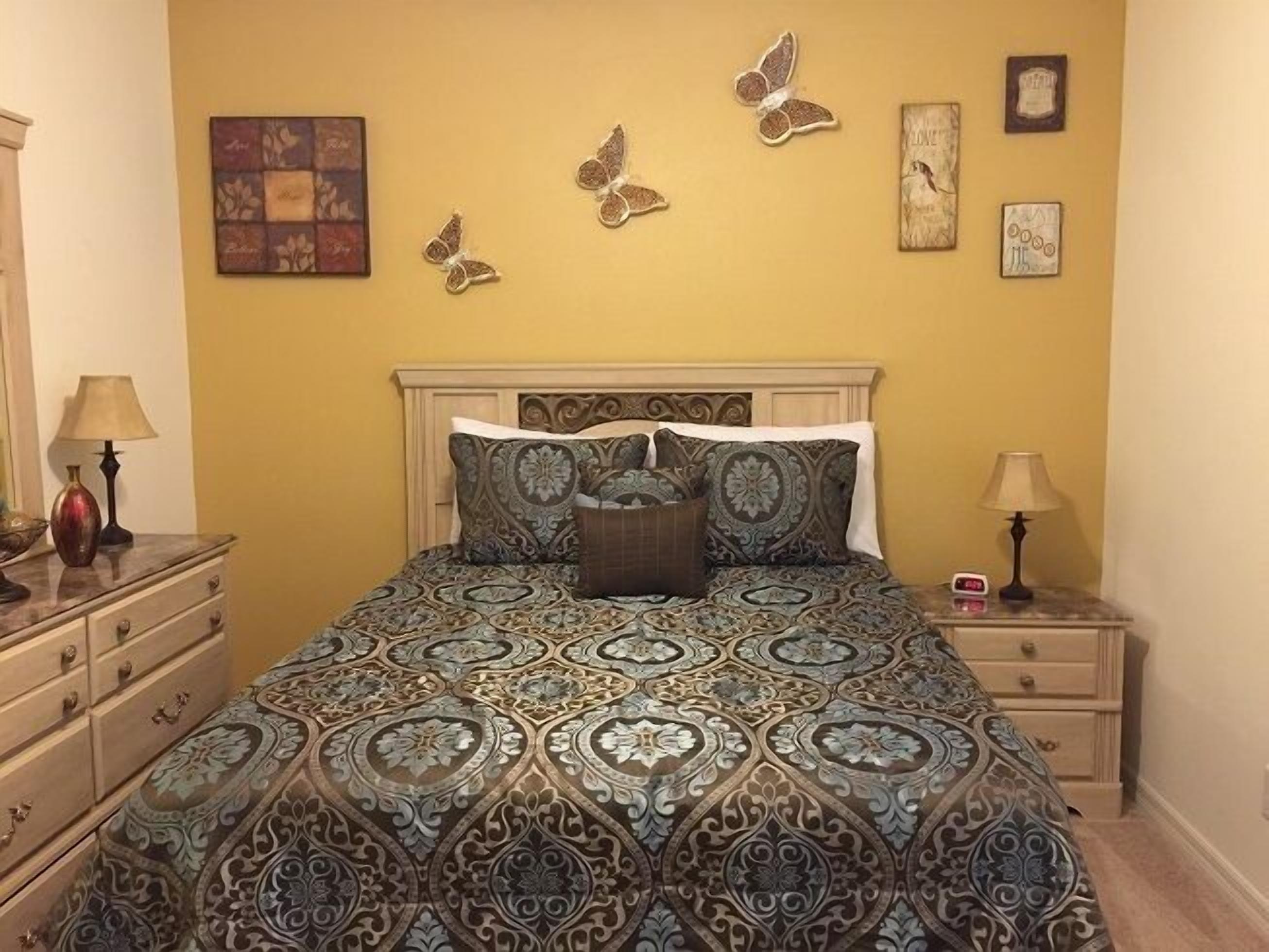 Ein Schlafzimmer mit einem gestreiften Bettüberwurf, einem Holzkopfende und Wanddekorationen, darunter Schmetterlingsmotive.