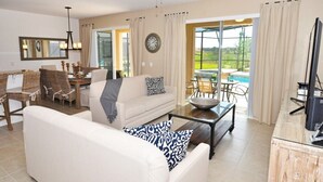 A living room with a white sofa, a coffee table, and a TV on a wooden stand. There is a dining area with a table and chairs, and a large window with curtains. - Solterra Resort IHR 3048 (Davenport)