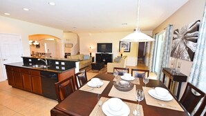 Un comedor amplio con una mesa grande dispuesta para cuatro personas, una cocina equipada con lavavajillas, y una sala de estar con sofá y televisor.