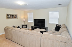 A living room with a beige sectional sofa, a flat-screen TV, and two windows with blinds.