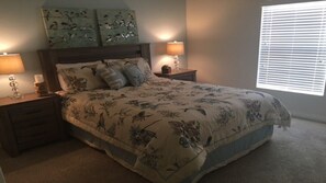 Quarto com colcha florida, duas luminárias de cabeceira, mesa de cabeceira e janela com persianas.