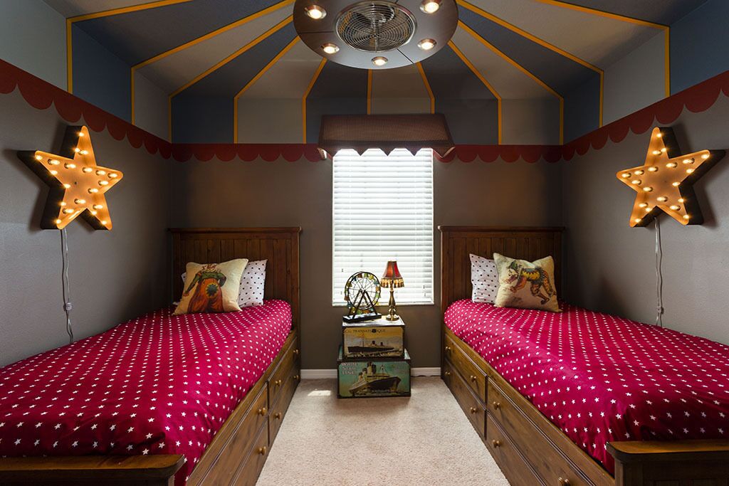 Habitación con dos camas, cada una con cubrecama rojo y decoración de estrellas en la pared. Cuenta con un ventilador de techo, una ventana con persianas y una pequeña mesa con una lámpara y un globo terráqueo.