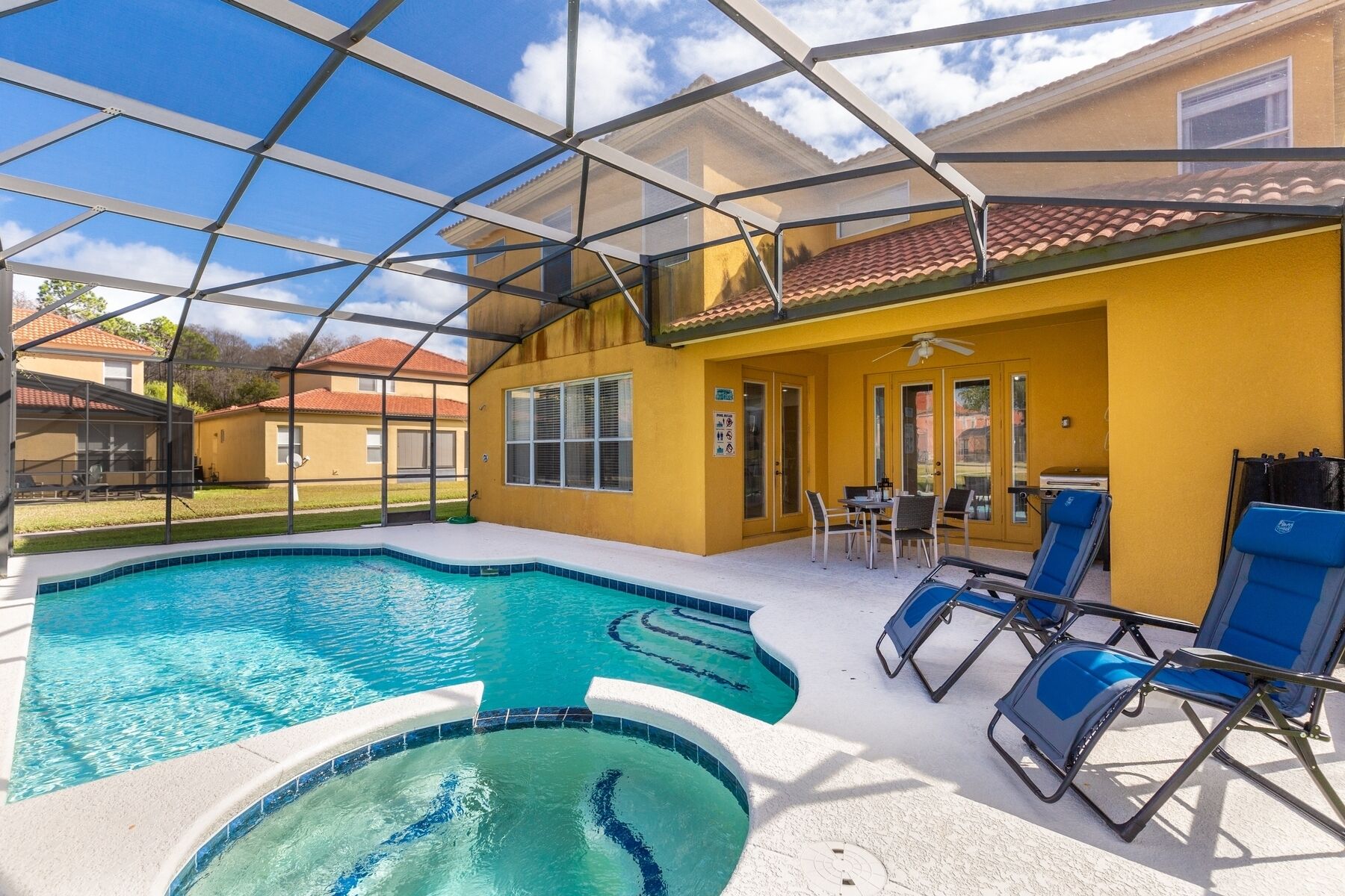 A swimming pool with a glass enclosure, a patio area with chairs, and a building with yellow walls and a red-tiled roof.