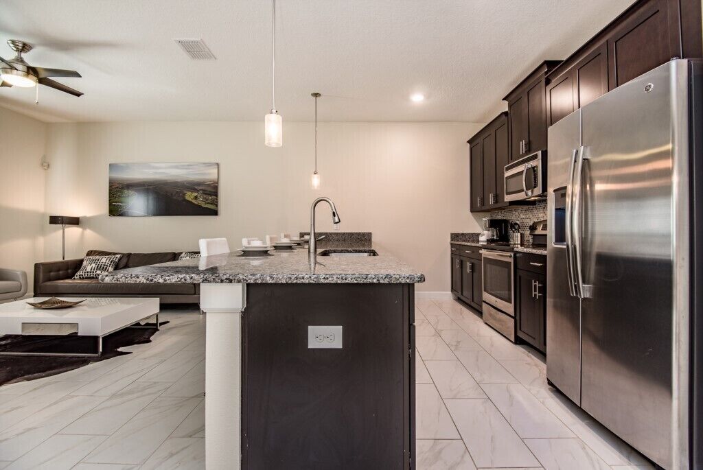 A modern kitchen with dark cabinets, stainless steel appliances, a granite countertop island, and a dining area with a sofa and coffee table.