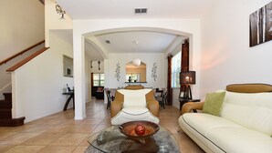 A spacious living room with a glass coffee table, a white sofa, and a dining area in the background.