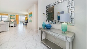 Un salon moderne avec un sol en marbre, une console ornée de vases décoratifs, un grand miroir à cadre géométrique et, au fond, un coin repas.