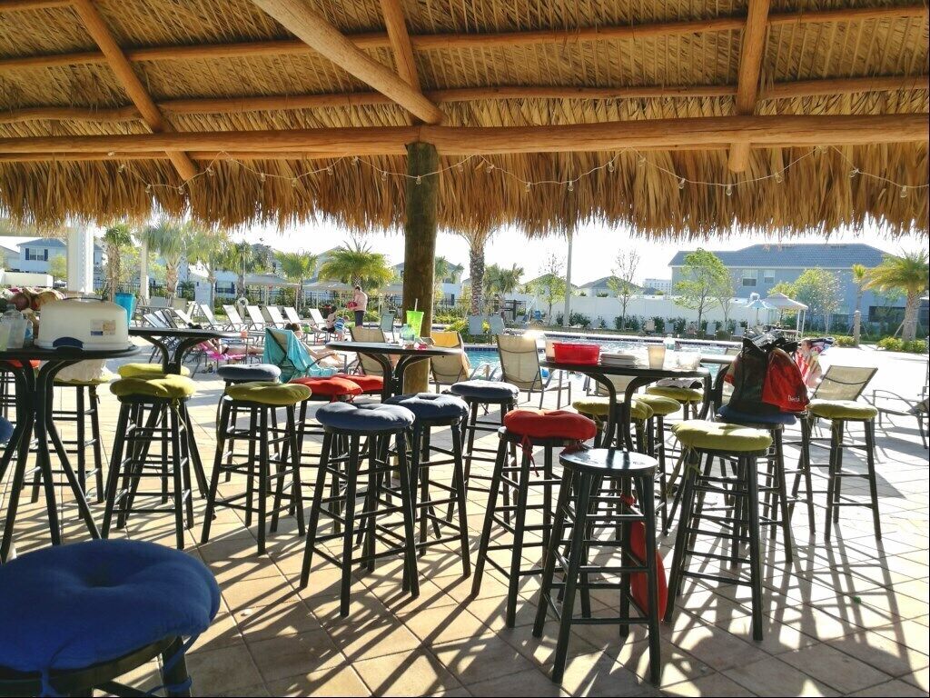 An outdoor seating area with multiple high stools and tables under a thatched roof, overlooking a pool and a residential area.