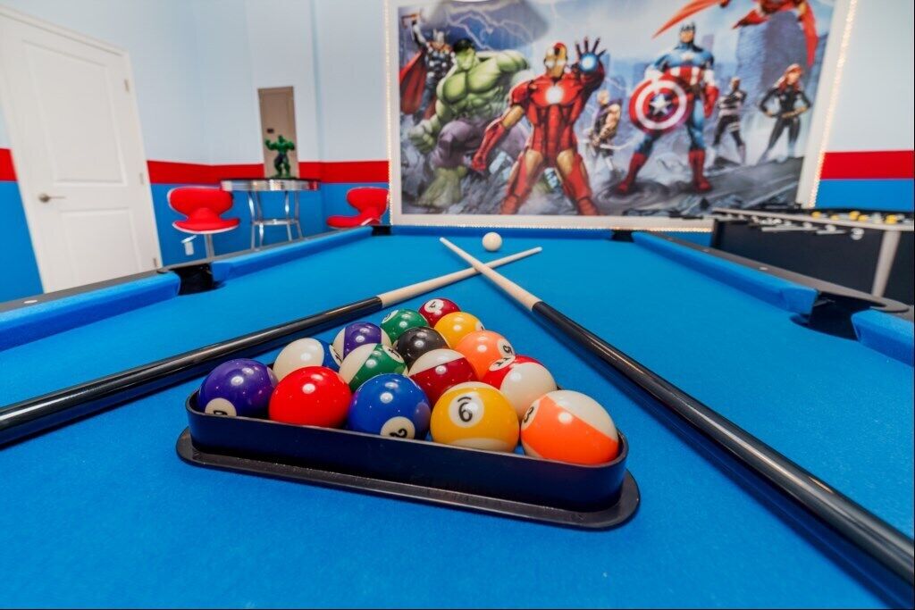 A pool table with colorful balls and a cue stick, a red chair, and a superhero-themed wall mural.