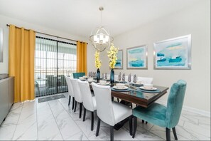 A dining area with a marble floor, a glass partition, yellow curtains, white chairs, a teal upholstered chair, a wooden table, and a modern chandelier.
