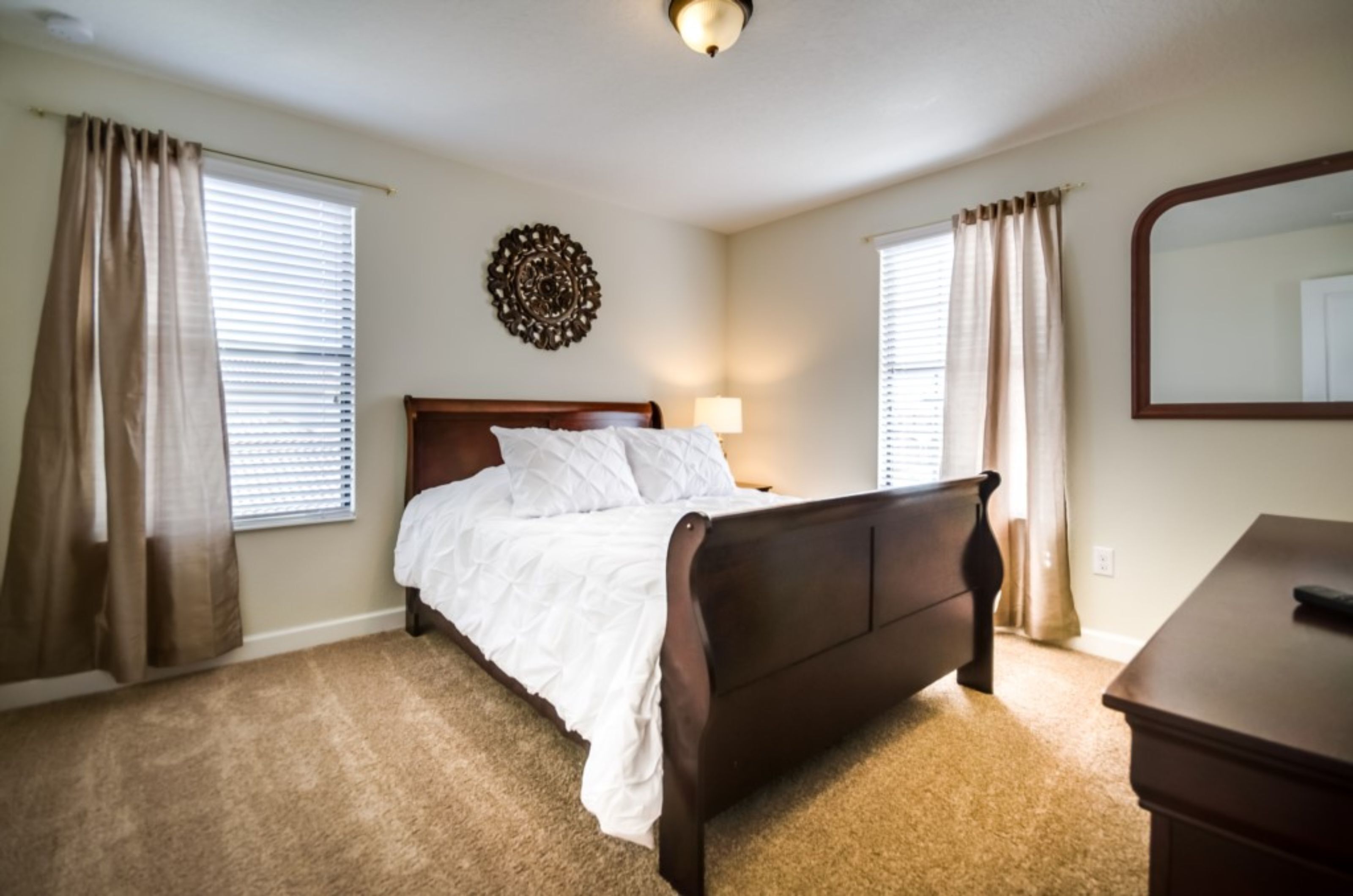 Un dormitorio con cama de madera, cortinas color beige, un espejo montado en la pared y piso cubierto de alfombra.