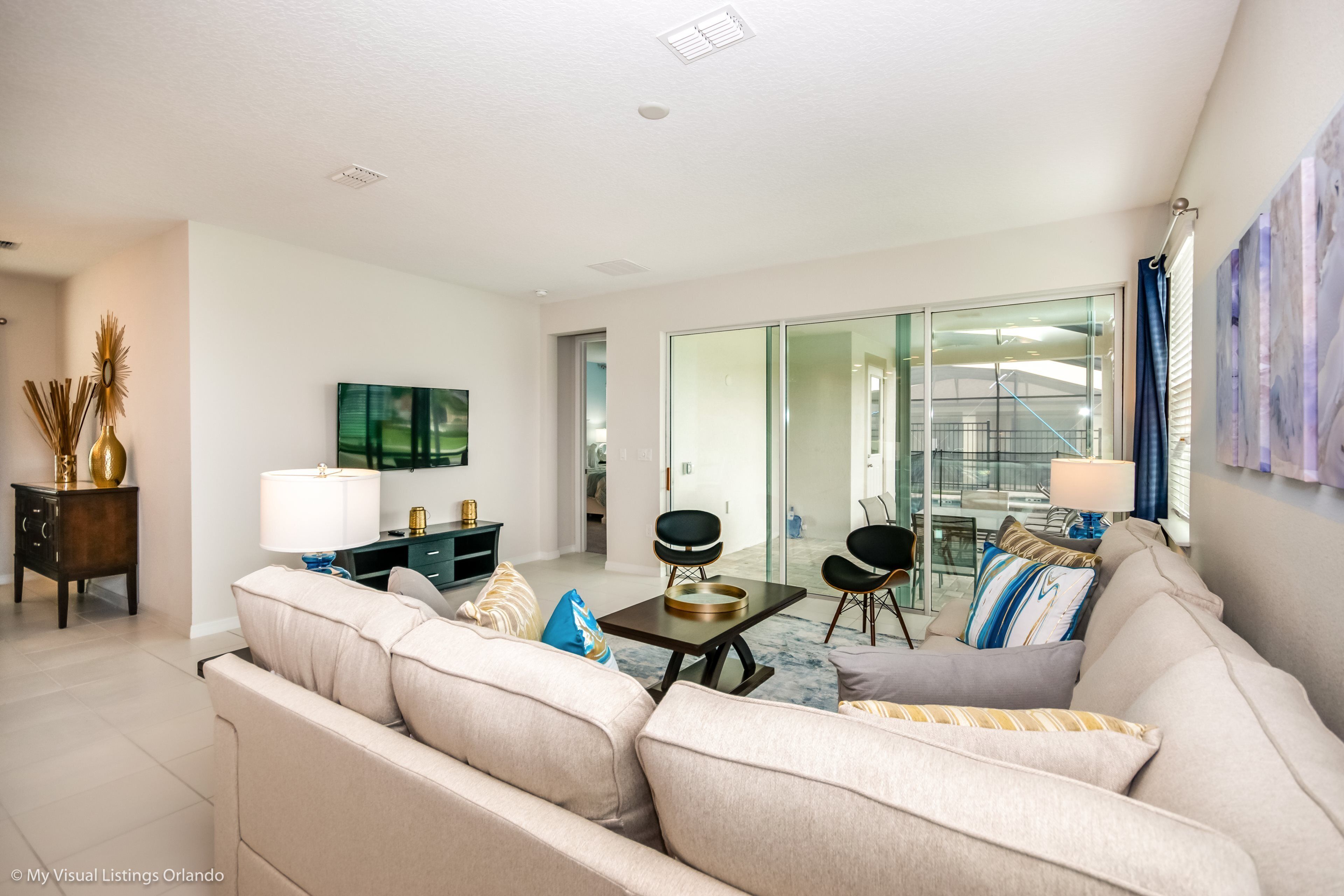 A modern living room with a beige sofa, a coffee table, and a TV. There is a glass door leading to a balcony.