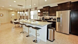 A modern kitchen with dark wooden cabinets, stainless steel appliances, a central island with bar stools, pendant lighting, and a large window providing natural light. - Luxury 5 Bedroom Private Pool IHR 1019 (Kissimmee)