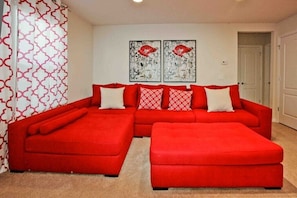 Um sofá modular vermelho com puf coordenado e almofadas decorativas estampadas, uma porta branca e uma parede com dois quadros de flores vermelhas.