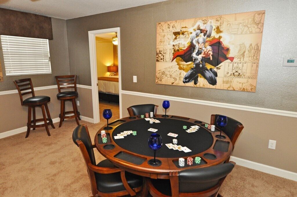 A room with a poker table set up for a game, a large superhero-themed artwork on the wall, and a doorway leading to another room.