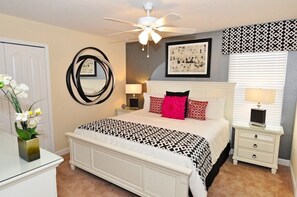 A bedroom with a white bed, a ceiling fan, a bedside table with a lamp, a mirror on the wall, and a framed picture above the bed.